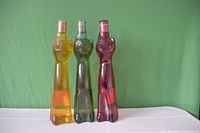 Three cat-shaped Moselland Riesling wine bottles in amber-orange, green, and dark red, arranged side by side on a white surface against a green background.