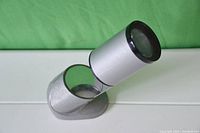 Silver cylindrical lens and angled mirror components photographed against a green background and white surface.