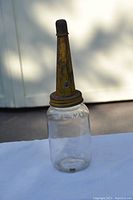 Clear glass jar with tall rusted metal spout and cap sitting on a white cloth, showing etched markings on the glass.