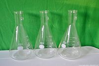 Five clear glass laboratory flasks against a green background showing size, shape and markings