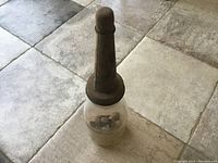 Clear view of the tall, transparent glass gasoline jar with rusted metal cap and conical nozzle, containing 7 extra metal tops inside.