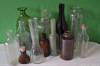 Photo showing 13 assorted glass bottles and jars of varying heights and colors on green background.