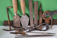 Photo of assorted old hand tools laid out on a green surface including trowels, rasps, pliers, punch, files, and a hex key.