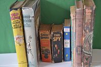 Photo showing all 8 books standing upright against a green background, spines and front covers visible, identifying titles and approximate condition.