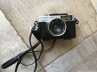 Front view of Ricoh 126-C camera showing lens, body and attached strap lying on stone tile floor.