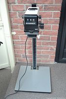 Full view of the enlarger standing on its metal base with power cord visible, against brick background.