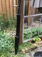 Tall, slender black metal garden seeder tool leaning against a glass window and wooden fence with plants surrounding it.