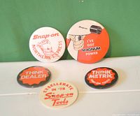 Five vintage Snap-On Tools buttons arranged on green and clear surfaces with various logos and slogans from 1970s-1980s.