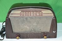 Front view of vintage brown Standard Broadcast radio with frequency dial, speaker grill, and control knobs showing wear and use marks.