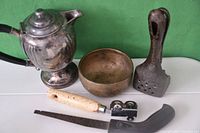 All items in the lot arranged on white surface with green background: tea pot, brass bowl, lemon squeezer, perforated tool, file, curved blade.