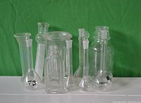 Photo showing 10 assorted clear glass lab containers including test tubes, flasks, and bottles with stoppers, one has chip on top.