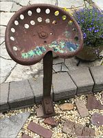 Front angle of the rusty vintage tractor seat with holes and green paint remnants on the seat surface, mounted on a metal base.