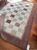 Full view of rectangular area rug on wooden floor, showing geometric square pattern with muted earth tones and dusty rose border.