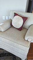 Front angle of beige upholstered armchair with decorative cushion showing fabric texture and arm design.