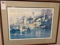 Framed artwork showing a harbor scene with sailboats and buildings along the water, framed in wood with a double mat.