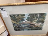 Framed watercolor painting showing the full scene of a gently curved bridge over water with trees around.