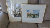 Two framed artwork pieces side by side on a couch, showing both cityscape prints with gold frames and white mats
