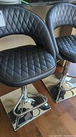 Side view of two black faux leather bar stools with diamond-quilted seat and backrest, chrome base with footrest and square bottom plate.