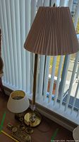 Tall brass floor lamp with beige pleated lampshade and 18-inch brass desk lamp with white shade, both standing on floor near blinds.