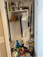 Photo showing various cleaning sprays, wooden and white plastic hangers, mop, hand brush, and cleaning cloths arranged inside a small closet space with a white shelf and rod.