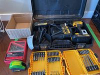 Open black carrying case showing the cordless drill, charger, two yellow bit holders with various bits, a red metal case with drill bits, and a tape measure.