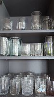 Shelves of assorted clear glassware showing different shapes of tumblers, beer mugs and drinking glasses with some having textured or patterned designs.