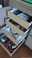 Drawer containing metal cutlery, white ceramic mugs, and stainless steel tools