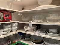 Shelved kitchenware including white French onion soup bowls with handles, stacked plates, clear glass bowls, and serving platters.