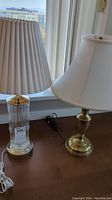 Two lamps side by side, glass chrome base lamp on the left, brass lamp on the right with fabric shades, placed on a wooden surface near window.