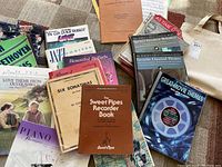 Photo showing a collection of music books including piano, recorder, vocal, and movie theme sheet music scattered and stacked on a surface.