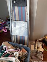 Aluminum stepladder leaning against the wall next to striped ironing board. Blue storage basket piled with assorted plastic coat hangers in pastel colors visible in foreground, part of plastic laundry baskets below.