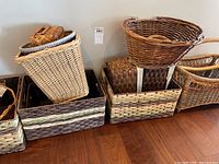 Photo showing an array of wicker and woven storage baskets of varying sizes and colors stacked against a wall on a wooden floor.