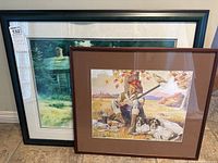 Two framed prints leaning against wall on tile floor; larger rustic cabin print behind smaller hunter scene print.