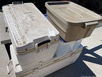 Top view showing two white Igloo coolers stacked and two storage bins, beige with lid and translucent with blue lid, stacked outdoors on concrete surface.