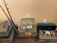 Wide view showing fishing rods leaning against the wall next to two tackle boxes, one beige and one dark green. Beige tackle box is open with fishing tackle inside.