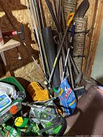 Photo shows a collection of fertilizers, spreader, rakes, shovels, brooms, and bags of fertilizer stacked against a wooden wall in a shed.