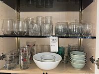 Shelf with various drinking glasses and bowls neatly arranged: clear stemless wine glasses, patterned short glasses, thick grey tumblers, tall nautical design glasses, white and green bowls.