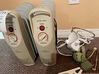 Photo showing two beige Air Works Mini Oil Heaters with power buttons, thermostat dials, and label markings. Also visible are two vintage desktop fans on the floor nearby.