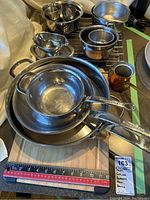 Photo showing the set of stainless steel pans stacked, metal bowls, a copper pot, gravy boat, and bamboo cutting boards with measuring tape for size reference.