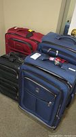 Four different sized luggage suitcases arranged on the floor, showing red, blue, and black colors with front pockets, handles, and wheels.