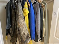 Men's outerwear hanging on rack showing camouflage jacket and pants, yellow raincoat, blue jacket, gray sweater, beige sweater vest, flotation jacket.