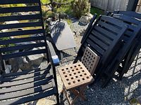 Four black painted wooden folding chairs alongside two natural wood lattice top folding tables and one gray resin and metal folding side table in outdoor setting on gravel.