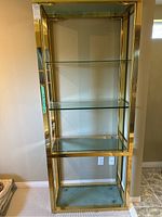 Full tall metal and glass shelving unit with brass or gold-tone frame and four clear glass shelves standing against a wall.