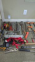 Wide view of the full assortment of hand tools laid out on a carpet, showing various wrenches, pliers, saws, hammers, clamps, and other hand tools.