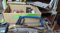 Various tools and hardware showing hand saw, hacksaw, hammer, and adjustable wrench in front of box with assorted nails and hardware.