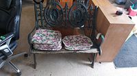 Front view of the bench with stacked floral seat pads placed on the weathered wooden seat, showing the ornate cast iron backrest and armrests.