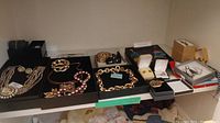 Wide shot showing multiple jewelry pieces laid out on black trays including necklaces, bracelets, brooches, watches, earrings, and rings