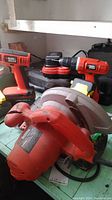 Two Black and Decker cordless drills in orange and black along with a circular saw on a table. Battery charger and batteries visible in background. Worn condition.