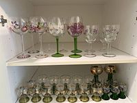 Two shelves of assorted glasses: top shelf contains hand-painted wine glasses with floral designs; bottom shelf has gold-accented shot glasses with green stems.