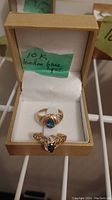 Two gold rings displayed in a jewelry box with a label stating '10K London blue topaz'. Both rings have topaz gemstones; the larger ring has a pear-shaped blue stone, the smaller ring has a round dark blue stone with detailed band.